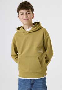 Niño con cabello corto y rizado, vestido con una sudadera amarilla mostaza y jeans azules, sonriendo levemente con las manos detrás de la espalda sobre un fondo blanco.