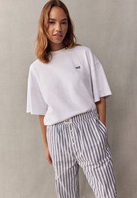 Bijela pamučna oversize majica s logotipom "self.", u kombinaciji sa sivim i bijelim prugastim hlačama koje imaju vezicu i kopčanje na gumbe.