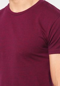 Homme portant un T-shirt bordeaux uni à manches courtes, montrant le haut du torse et le cou sur un fond blanc.