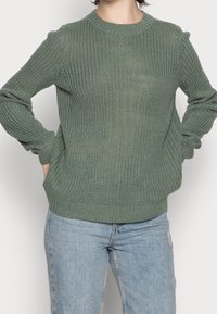 Grüner gestrickter Pullover mit strukturiertem Muster, rundem Halsauschnitt und gerippten Bündchen und Saum. Kombiniert mit hellblauen Jeans.