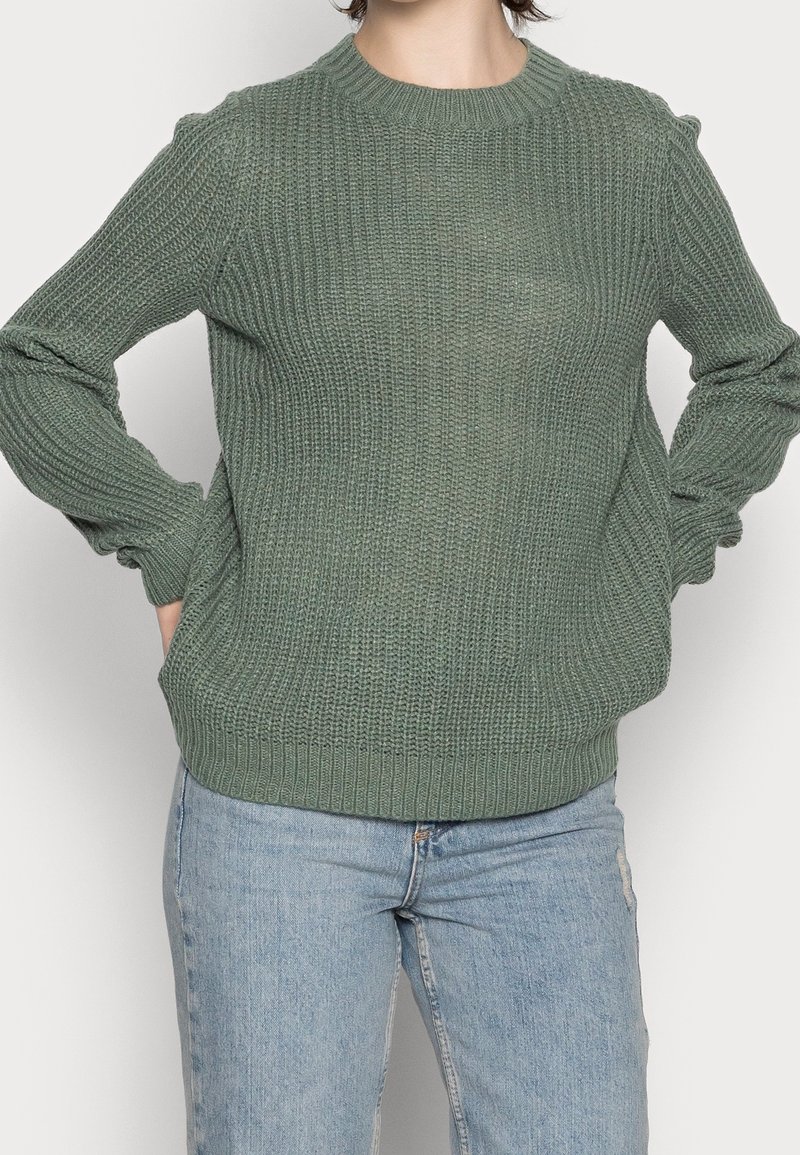 Grüner gestrickter Pullover mit strukturiertem Muster, rundem Halsauschnitt und gerippten Bündchen und Saum. Kombiniert mit hellblauen Jeans.