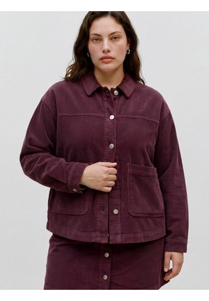 Femme aux cheveux bruns portant des boucles d'oreilles en créoles argentées et une veste en velours côtelé violet foncé boutonnée avec une jupe assortie, debout devant un fond uni.