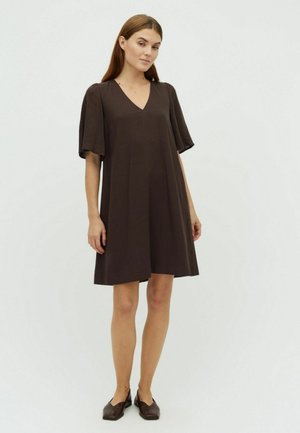 Frau mit langen braunen Haaren, die ein lockeres, kurzärmeliges dunkelbraunes Kleid und braune flache Schuhe trägt und vor einem schlichten hellen Hintergrund steht.