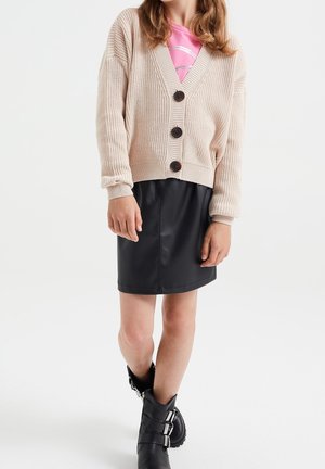 Dziewczyna w beżowym, zapinanym na guziki kardiganie na różowej bluzce, czarnej skórzanej spódnicy i czarnych botkach z klamrami, stojąca z nogami skrzyżowanymi na białym tle.