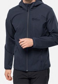 Chaqueta de softshell negra con cierre frontal con cremallera, capucha ajustable, bolsillos laterales y detalle de logo, que presenta un diseño ajustado y una textura suave.