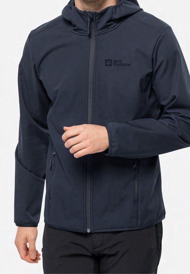 Chaqueta de softshell negra con cierre frontal con cremallera, capucha ajustable, bolsillos laterales y detalle de logo, que presenta un diseño ajustado y una textura suave.