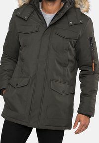 Zielony oliwkowy płaszcz zimowy z kapturem wyściełanym sztucznym futrem, wieloma kieszeniami, zapięciem na zamek i zatrzaski oraz teksturowanym wykończeniem tkaniny.