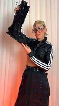 Botas altas até ao joelho em preto com atacadores, em couro texturizado, com laço lateral e sola plana. Modelo a usar uma jaqueta e saia pretas, segurando a bota.