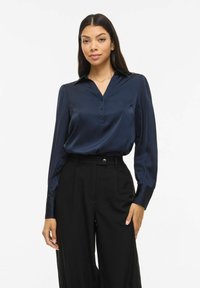 Tumši zila satīna blūze ar apkakli, pogu priekšpusi un garām piedurknēm; kopā ar melnām augsta vidukļa biksēm ar pogu detaļu.