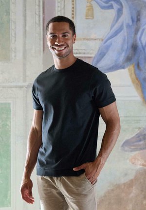 Black cotton t-shirt with a crew neck and short sleeves, fitted silhouette, paired with light beige pants, standing against a painted backdrop.