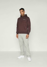 Hanorac maro cu șnururi, buzunar frontal și un mic logo. Asortat cu pantalonii de trening gri deschis și adidași albi. Design simplu, potrivire casual.