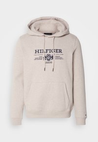 Sudadera de color beige claro con capucha ajustable, bolsillo frontal y estampado de logo en azul marino. Presenta puños y dobladillo acanalados. Tejido suave y texturizado.