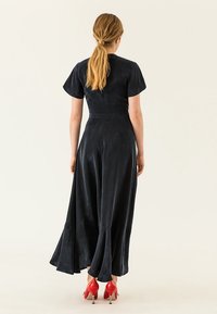 Černé semišové maxi šaty s krátkými rukávy, těsným korzetem a rozšířeným lemem. Modelka má na sobě červené vysoké podpatky.