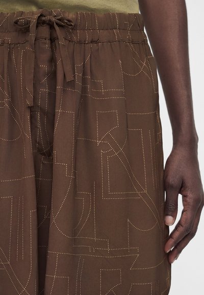 Σκούρο καφέ παντελόνια με ελαστική μέση και κορδόνι, με ανοιχτά πουά αφηρημένα γραμμικά μοτίβα, φορεμένα από άτομο με σκουρόχρωμο δέρμα.