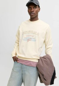 Lys gul crew-neck sweatshirt med frontgrafik; lagt over en pink skjorte. Båret sammen med lyseblå jeans og holder en brun jakke.