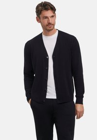 Cardigan blu scuro realizzato in tessuto morbido, con scollatura a V, maniche lunghe e sei bottoni frontali. Indossato sopra una camicia bianca.