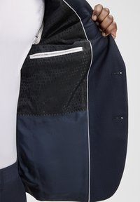 Marineblauer Anzugsakko mit einem schwarzen gemusterten Futter, weiß eingefasstem Rand, Innentasche und zwei Außenknöpfen an den Ärmeln.