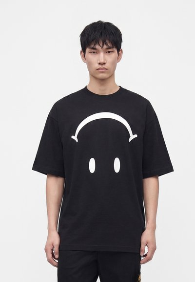 Jeune homme aux cheveux noirs courts portant un t-shirt noir oversized avec un grand smiley blanc à l'envers sur le devant.