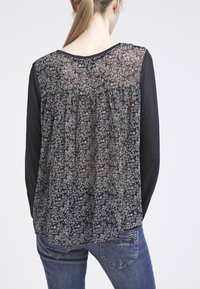 Zwarte lange mouwen top met een doorschijnende achterkant met een wit bloemenpatroon en geplooide stof, gecombineerd met blauwe denim jeans.