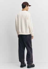 Crèmekleurig sweatshirt met een relaxte pasvorm en een ronde hals, gecombineerd met donkerpaarse wijde jeans. Model draagt een bruine pet en zwarte laarzen.