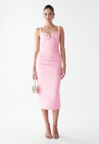 Robe rose ajustée avec un décolleté en cœur, détails froncés au niveau de la poitrine et des côtés, et longueur midi droite. Modèle tenant un petit sac.