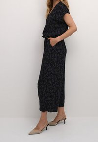 Mono negro con flores, de manga corta y escote en V. Presenta un ajuste relajado, bolsillos laterales y una textura estampada. Combinado con tacones beige.