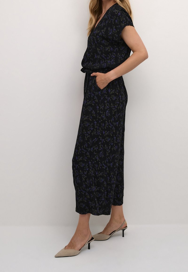 Mono negro con flores, de manga corta y escote en V. Presenta un ajuste relajado, bolsillos laterales y una textura estampada. Combinado con tacones beige.