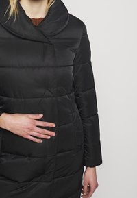 Abrigo acolchado negro con un cuello ancho y doblado, textura acolchada y un bolsillo lateral, hecho de una tela lisa y brillante.