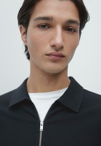 Czarna koszula na zamek błyskawiczny z kołnierzykiem, wyposażona w srebrny zamek błyskawiczny i noszona na białej koszulce. Gładka faktura tkaniny, minimalistyczny design.