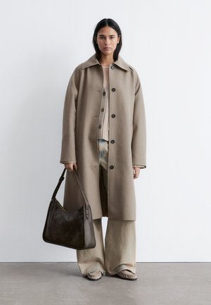 Woman wearing a beige long coat over a cardigan and wide pants, holding a dark brown handbag, standing against a white wall.