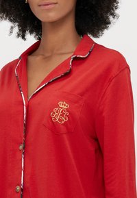 Tricou de pijama roșu dintr-un material ușor, cu guler. Include o buzunar pe piept cu broderie aurie și un tiv colorat de-a lungul gulerului.