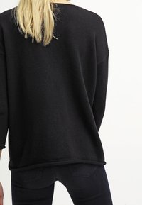 Pull en tricot noir avec une coupe ample et oversize, présentant un large col et des épaules tombantes, associé à un jean foncé.