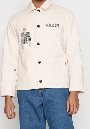 Beige skjortejakke med sort skeletgrafik på venstre bryst og teksten "VOLCOM" over brystlommen, båret med blå jeans.