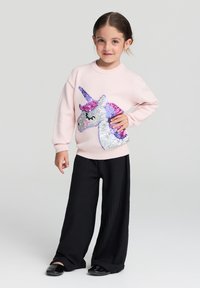 Ragazzina in piedi con la mano sui fianchi, che indossa un maglione rosa con un unicorno di paillettes e pantaloni larghi neri, su uno sfondo semplice.