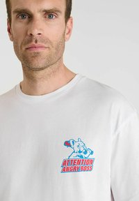 Man met kort bruin haar en een lichte baard, gekleed in een wit shirt met een blauw en rood grafisch ontwerp van een hond die een knuppel vasthoudt en de tekst "Let op, Boze Baas."