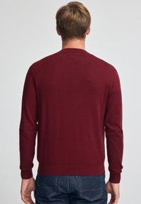 Maglione color bordeaux con maniche lunghe e scollo rotondo. Tessuto morbido, polsini e orlo a coste. Indossato sopra jeans di denim scuro.