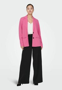 Růžový blazer s klopou, jedním knoflíkem, dvojicí předních kapes, nošený přes bílé tričko a černé široké kalhoty. Hladký materiál.
