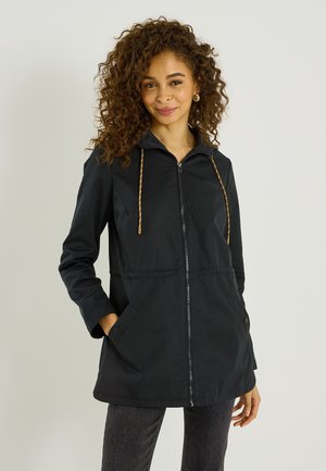 Femme aux cheveux bouclés portant une veste noire zippée avec cordons de serrage et poches avant, debout devant un fond clair uni.