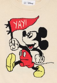 Crèmekleurige gebreide trui met een geborduurde Mickey Mouse die een rode vlag vasthoudt met "YAY!" in het wit. Gele schoenen en zwarte accenten maken het ontwerp compleet.