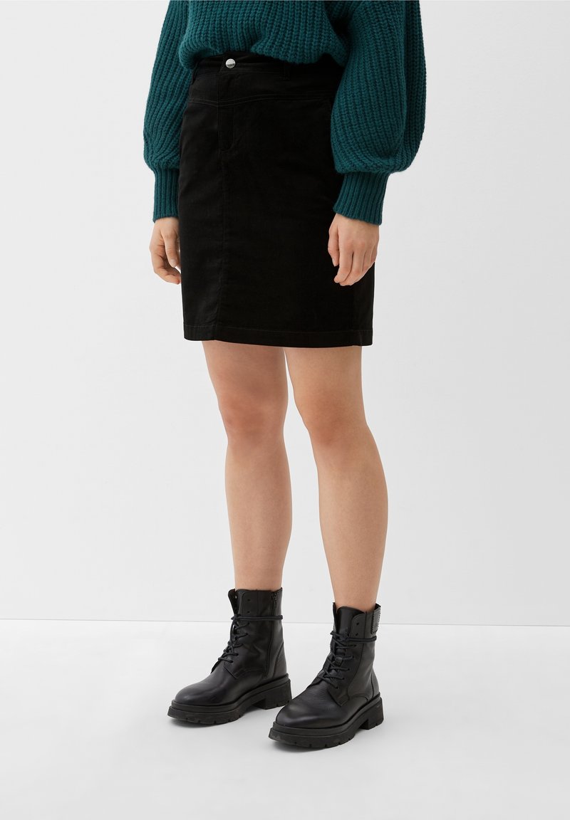Femme portant une mini-jupe noire, un pull en maille épaisse bleu sarcelle et des bottes de combat noires à lacets, debout sur un sol et un mur blancs.