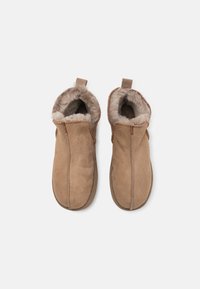 Beige semskede tøfler med en fluffy fòr, lukket design og teksturert søm langs kantene. Har bakre trekkfliker for enkel påkledning.