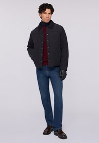 Svart vadderad jacka med tryckknappar, i kombination med en vinröd tröja och mörkblå jeans. Modellen bär svarta handskar och bruna skor.