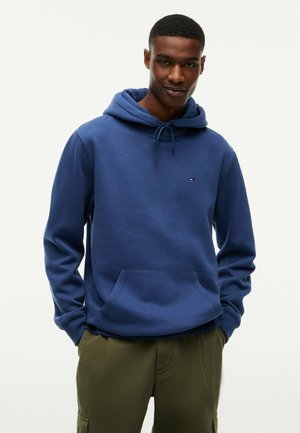 Mann trägt einen blauen Kapuzenpullover mit Fronttasche und olivgrünen Hosen und steht vor einem einfarbigen hellen Hintergrund.