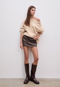 Pull en maille beige oversize, mini-jupe en cuir marron et bottes marron montant jusqu'aux genoux avec des lanières. Design simple et palette de couleurs chaudes.
