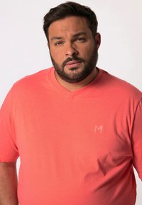 Korálové tričko s výstřihem do V vyrobené z měkkého materiálu s hladkou texturou, s jemným logem "M" v horním levém rohu.
