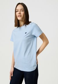 Jasnoniebieski bawełniany t-shirt z okrągłym dekoltem i krótkimi rękawami. Posiada małe ciemne logo po lewej stronie klatki piersiowej. Prosty fason, luźny krój.