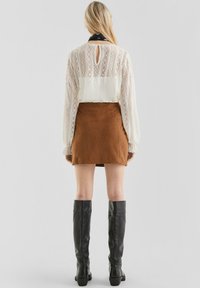 Donna che indossa un top bianco in pizzo a maniche lunghe, una mini gonna in suede marrone, una sciarpa nera al collo e stivali in pelle neri fino al ginocchio, di spalle.