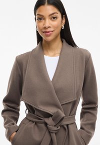 Abrigo envolvente marrón con cinturón y un gran cuello, hecho de una tela suave, con bolsillos laterales y una textura lisa. Combinado con una blusa blanca.