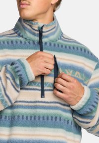 Fleeceový pulóver v modro-krémovej farbe s horizontálnymi pruhmi. Má zips na krku, elastické manžety a mäkkú, nadýchanú štruktúru.