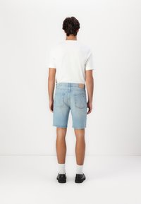 Shorts en denim bleu clair avec une coupe décontractée, dotés d'un patch au dos de la taille et de deux poches arrière. Portés avec un t-shirt blanc et des chaussures noires.